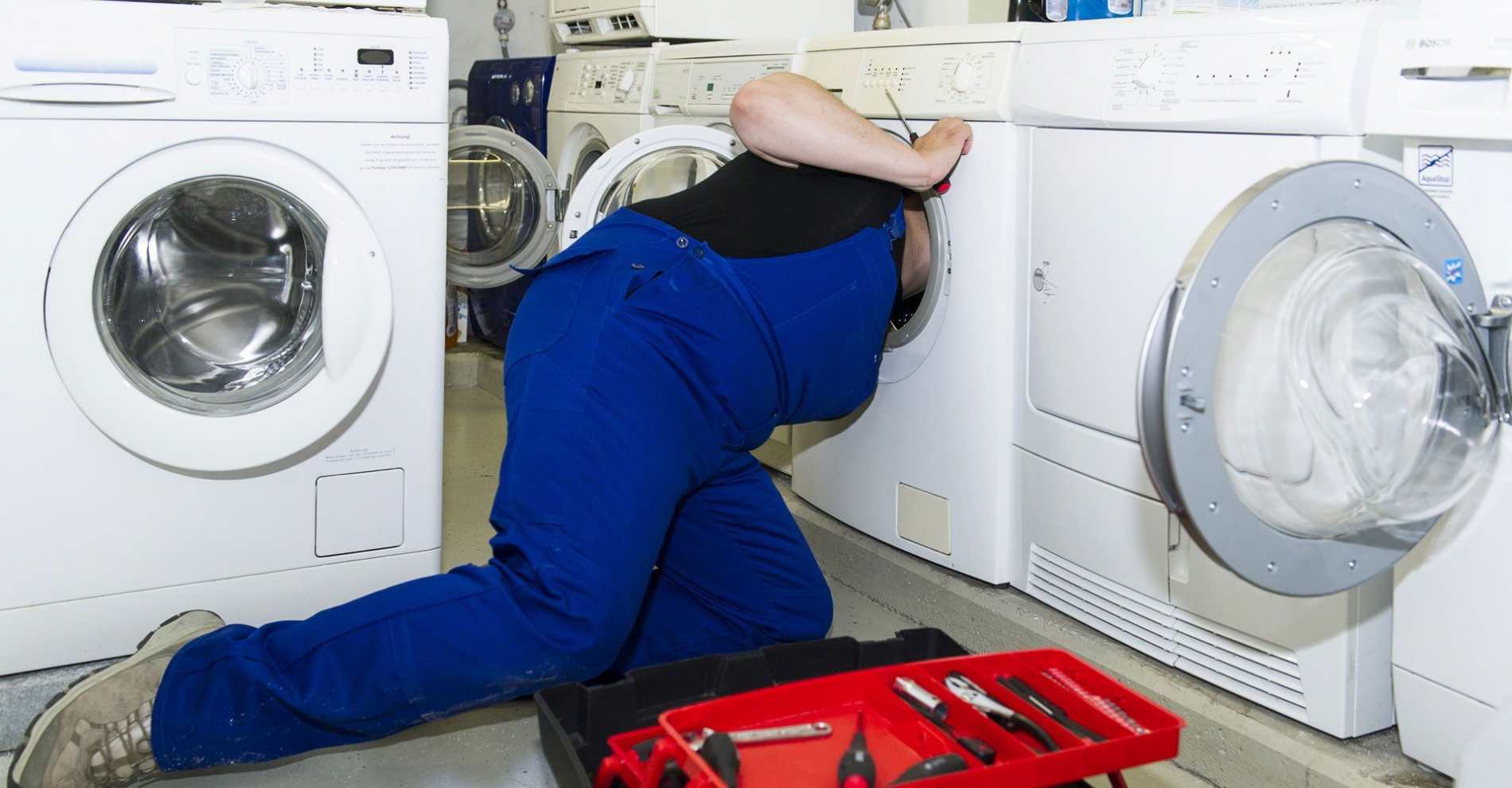 Appliance engineer kneeling beside a washing machine with a tool kit, carrying out a repair